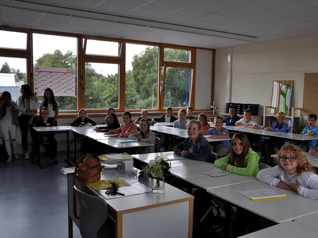 Kinder sitzen im Klassenraum, an der Seite stehen Erwachsene.